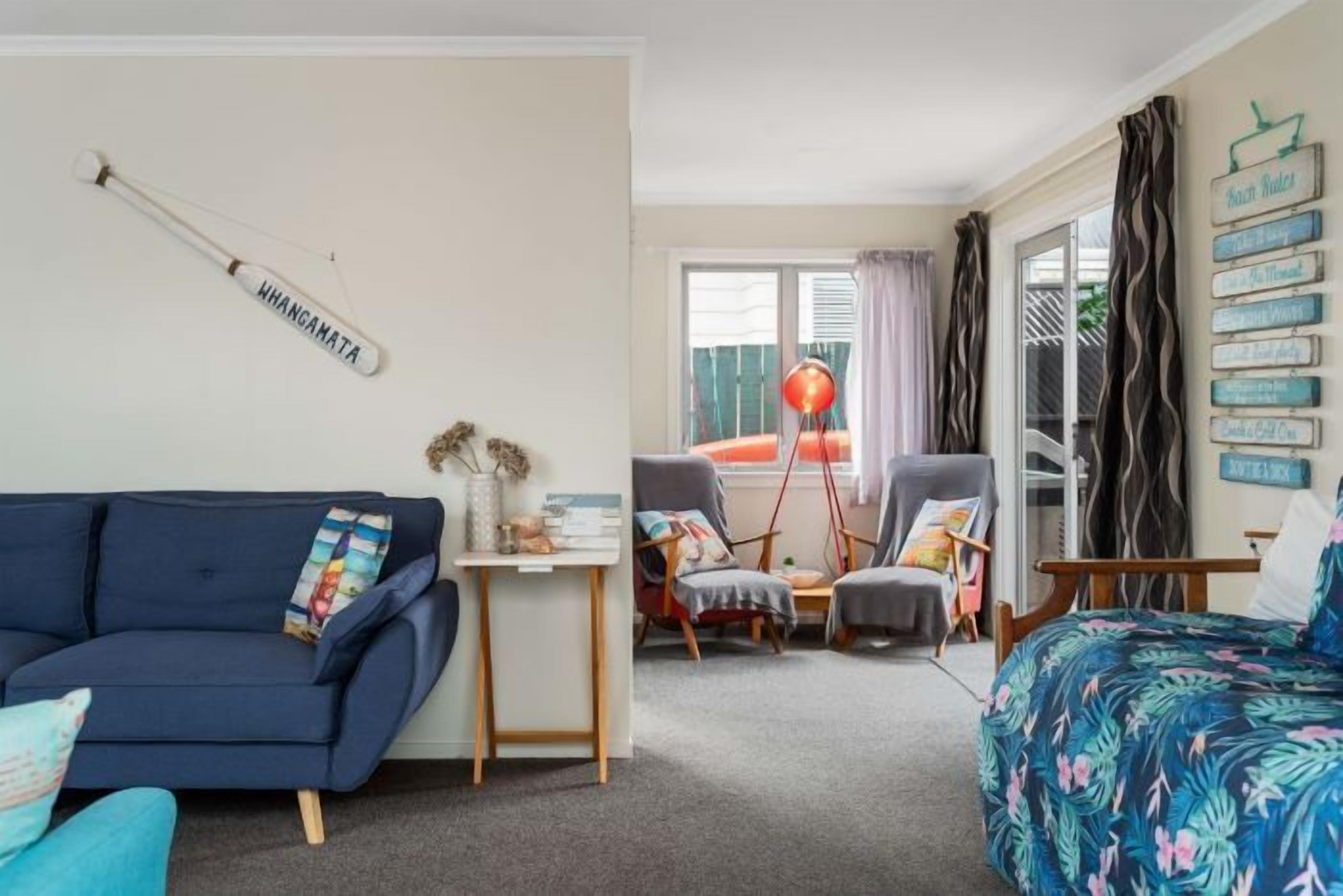 Ein gemütliches Zimmer mit einem blauen Sofa, einem kleinen Tisch mit Dekoration, zwei Sesseln, einem Bett mit floralem Überwurf und einem Fenster mit Vorhängen.