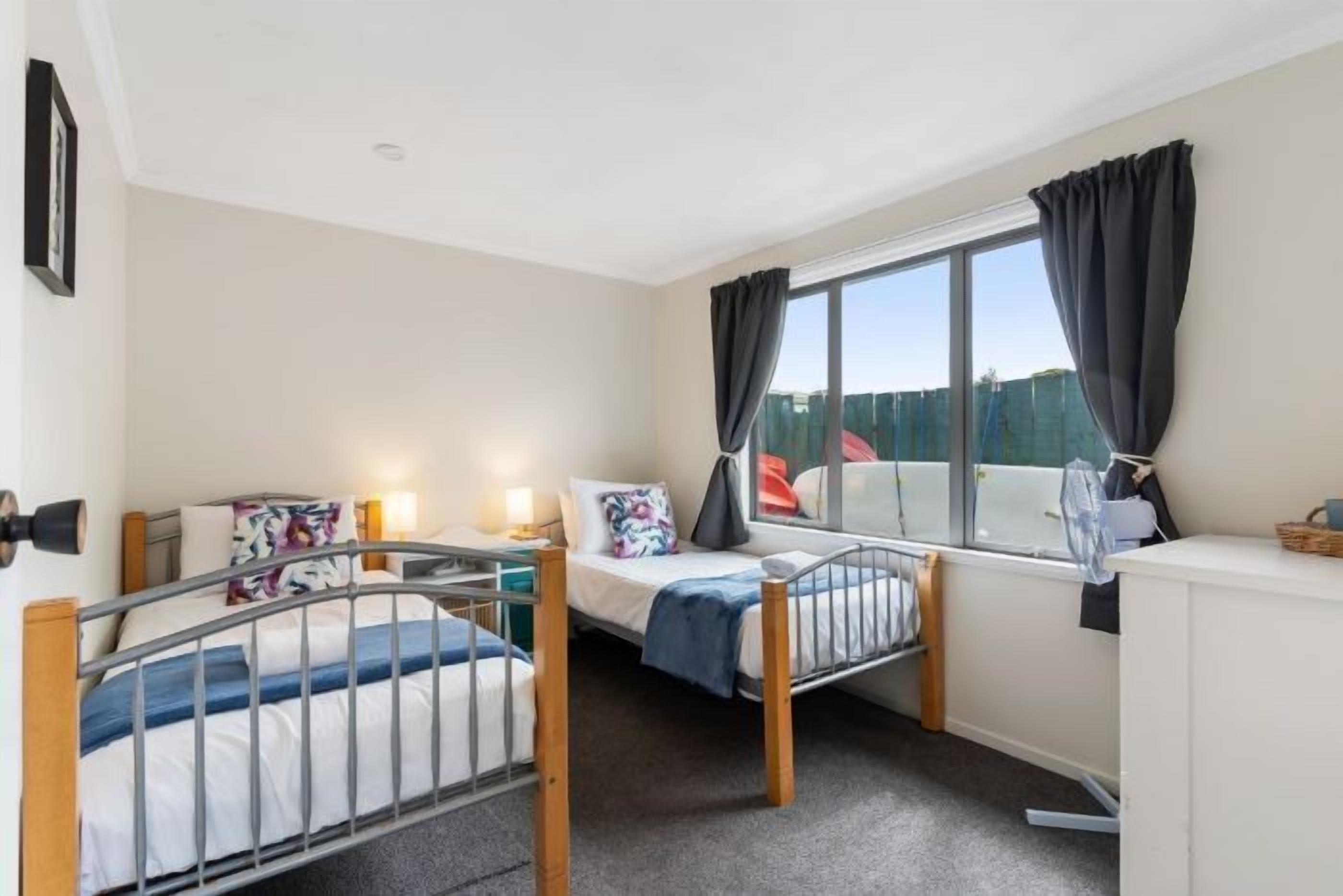 A small, well-lit room with two single beds, a desk, and a large window with curtains.