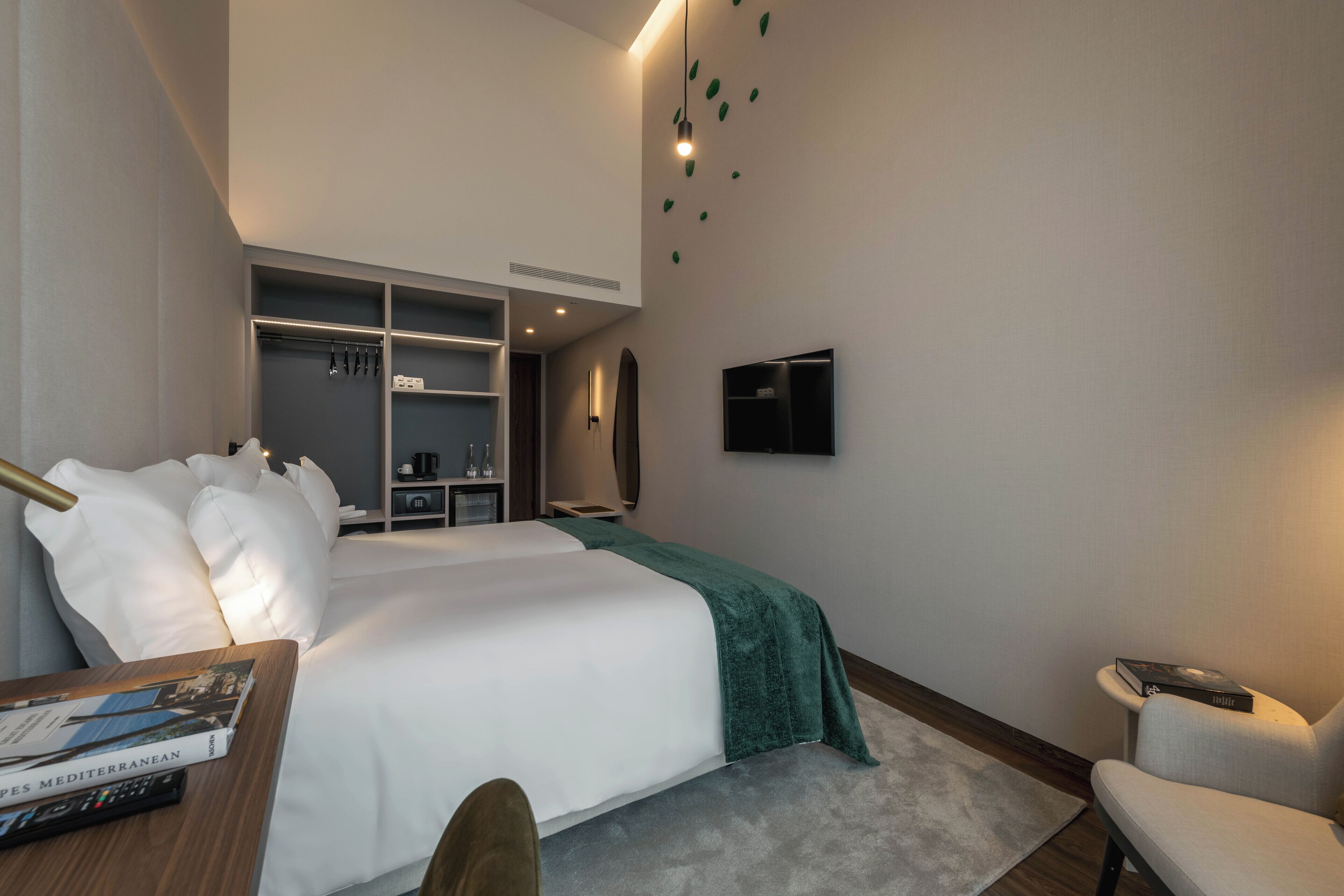 A modern hotel room with a large bed, a desk with a book, a chair, a TV, and a walk-in closet.