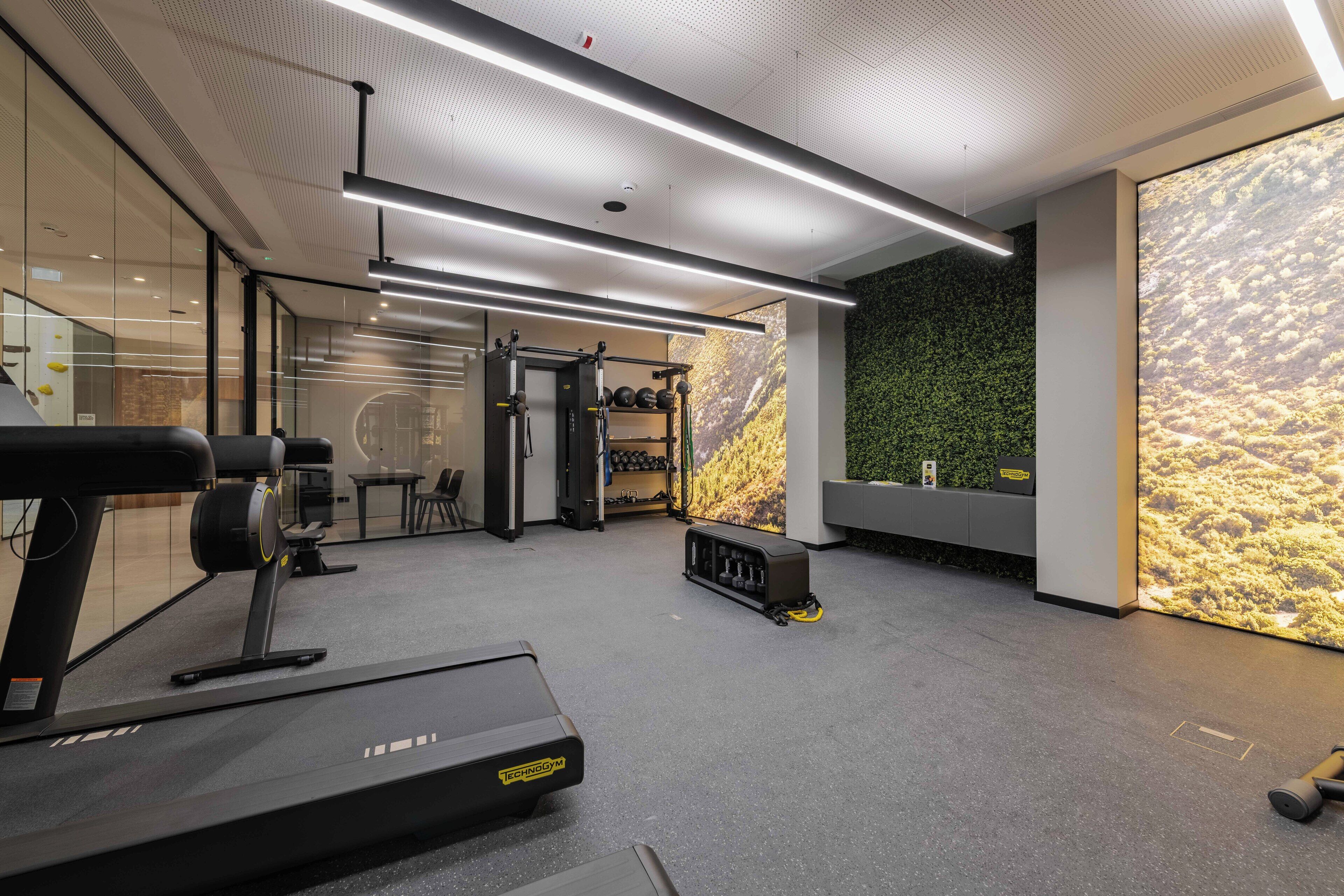 A modern gym with a treadmill, weight machines, and a green wall with a nature mural.