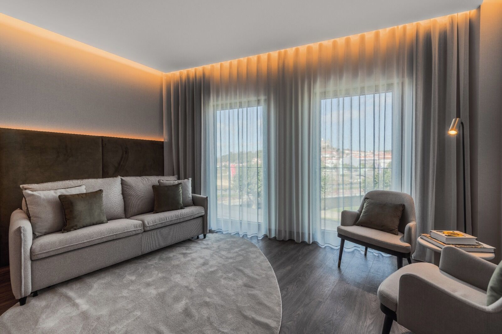 Ein modernes Hotelzimmer mit einer Couch, einem Sessel und großen Fenstern mit Vorhängen.