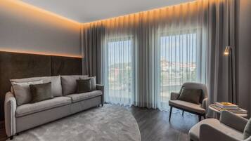 A modern hotel room with a sofa, armchair, and large windows with curtains.