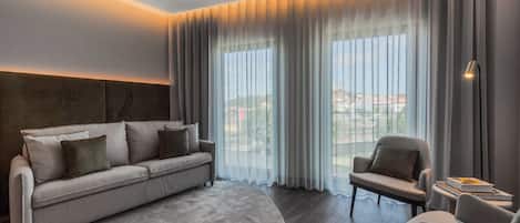 Ein modernes Hotelzimmer mit einer Couch, einem Sessel und großen Fenstern mit Vorhängen.