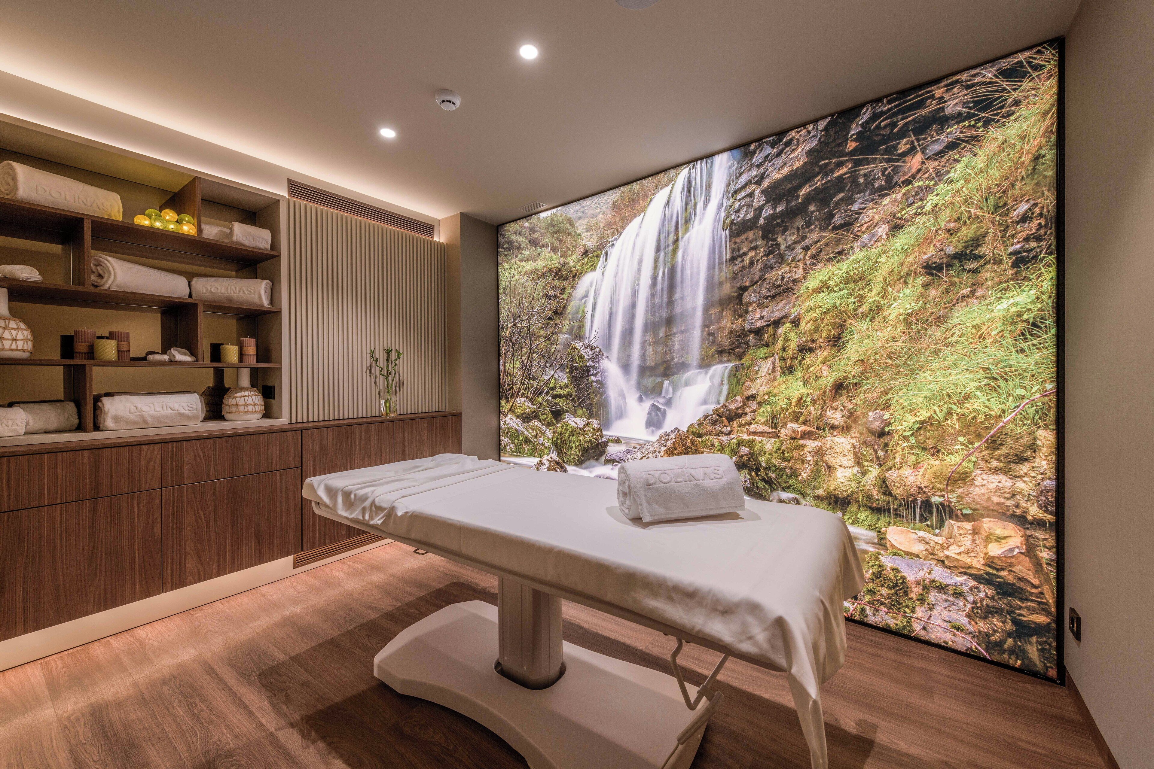 A spa room with a wooden cabinet, a massage table, and a large waterfall mural.
