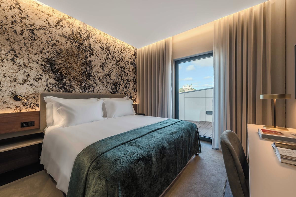 Ein modernes Hotelzimmer mit einem großen Bett, einem Schreibtisch mit Lampe und Blick auf die Stadt durch das Fenster.