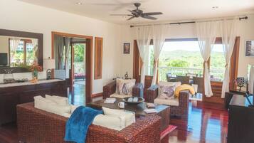 A spacious living room with wicker furniture, a ceiling fan, and large windows with curtains.