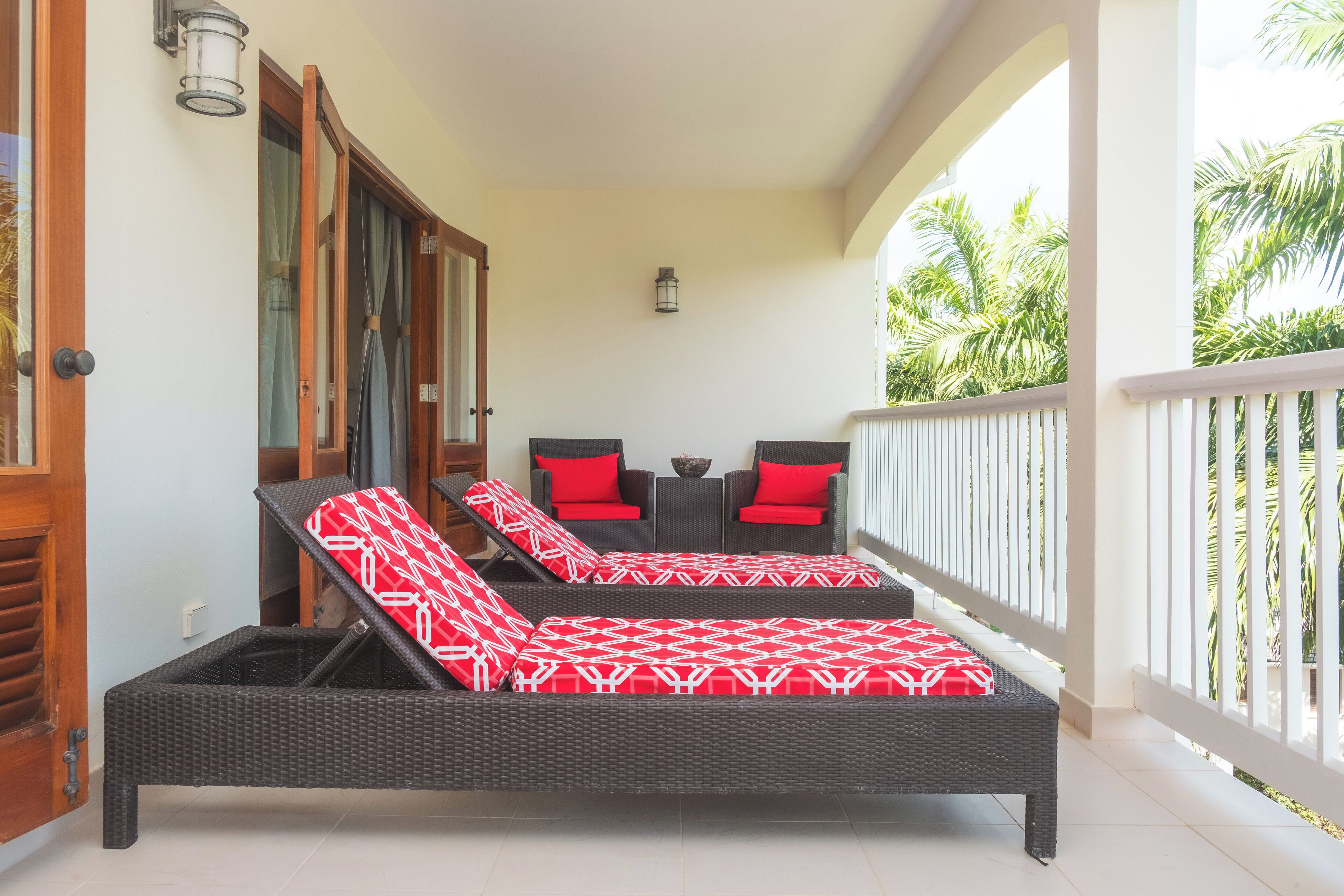 A balcony with wicker lounge chairs and red cushions, a small table, and a lantern.