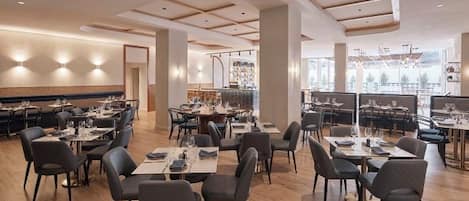 A spacious restaurant with multiple dining tables and chairs, a modern ceiling design, and large windows allowing natural light.