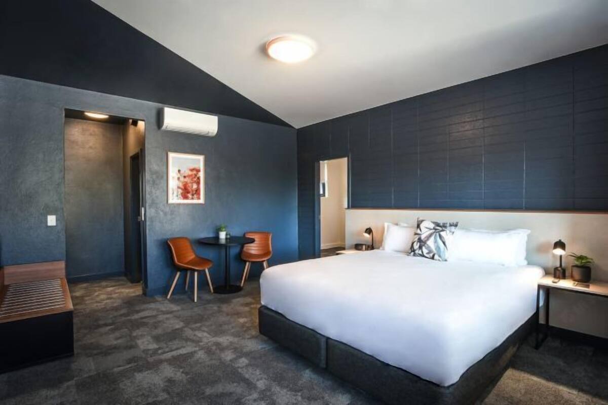 Moderni hotellihuone, jossa on suuri sänky, työpöytä tuolilla sekä pieni pöytä kahdella tuolilla.
