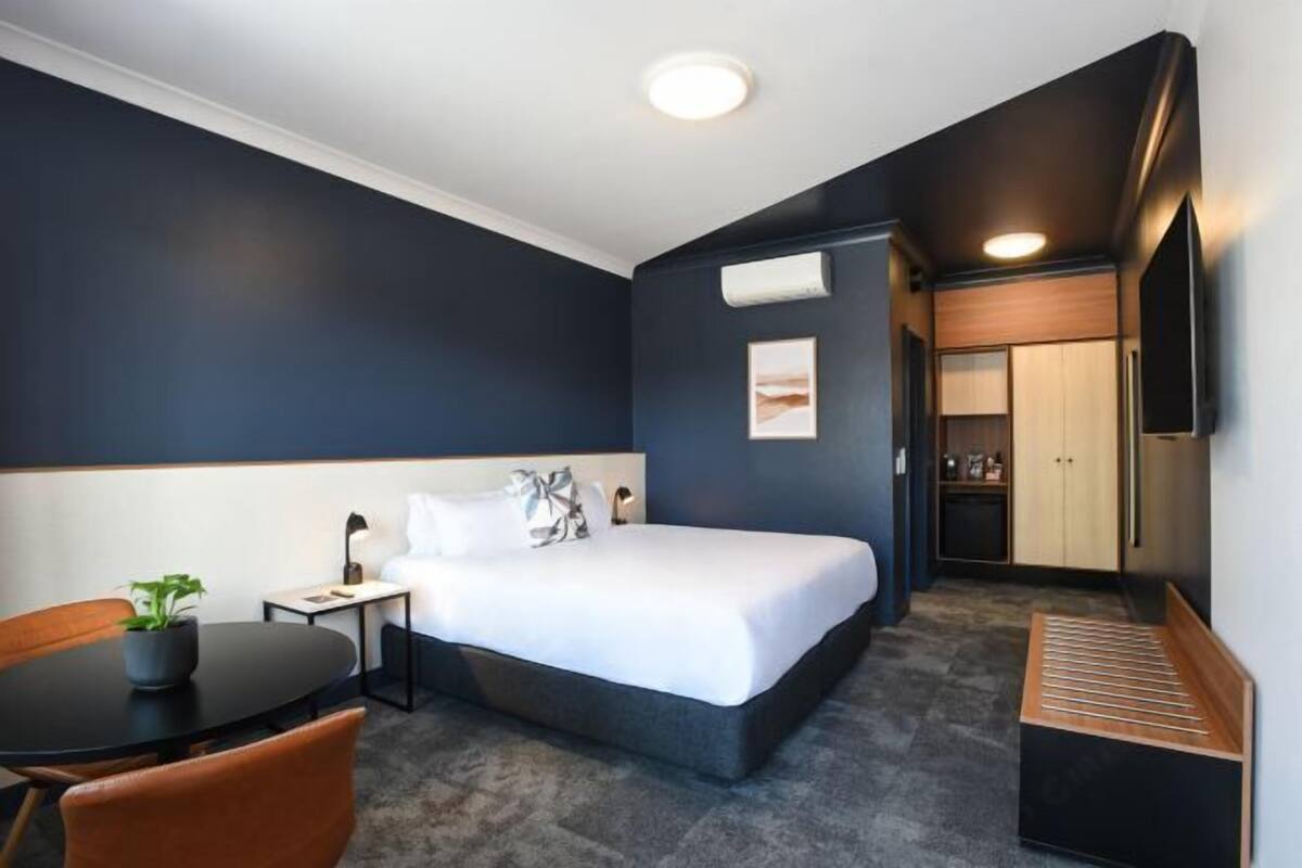 Hotellihuone, jossa on sänky, työpöytä, tuoli, pieni pöytä, jossa on kasvi, ja pieni keittotila.