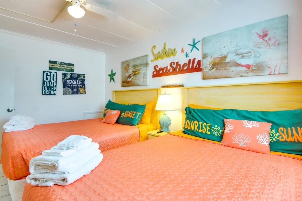 Ein Zimmer mit zwei Betten, jedes mit einer orangefarbenen Bettdecke und passenden Kissen. Auf der linken Seite des Bettes sind Handtücher gestapelt. Die Wände sind mit Kunstwerken und Schrift versehen, darunter 'Sunshine Seashells' und 'Go Jump in the Ocean'.