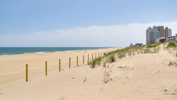 A sandy beach with a wooden fence and grass in the foreground, leading to a building complex in the background.
