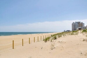 Una playa arenosa con una valla de madera y césped en primer plano, que conduce a un conjunto de edificios al fondo.
