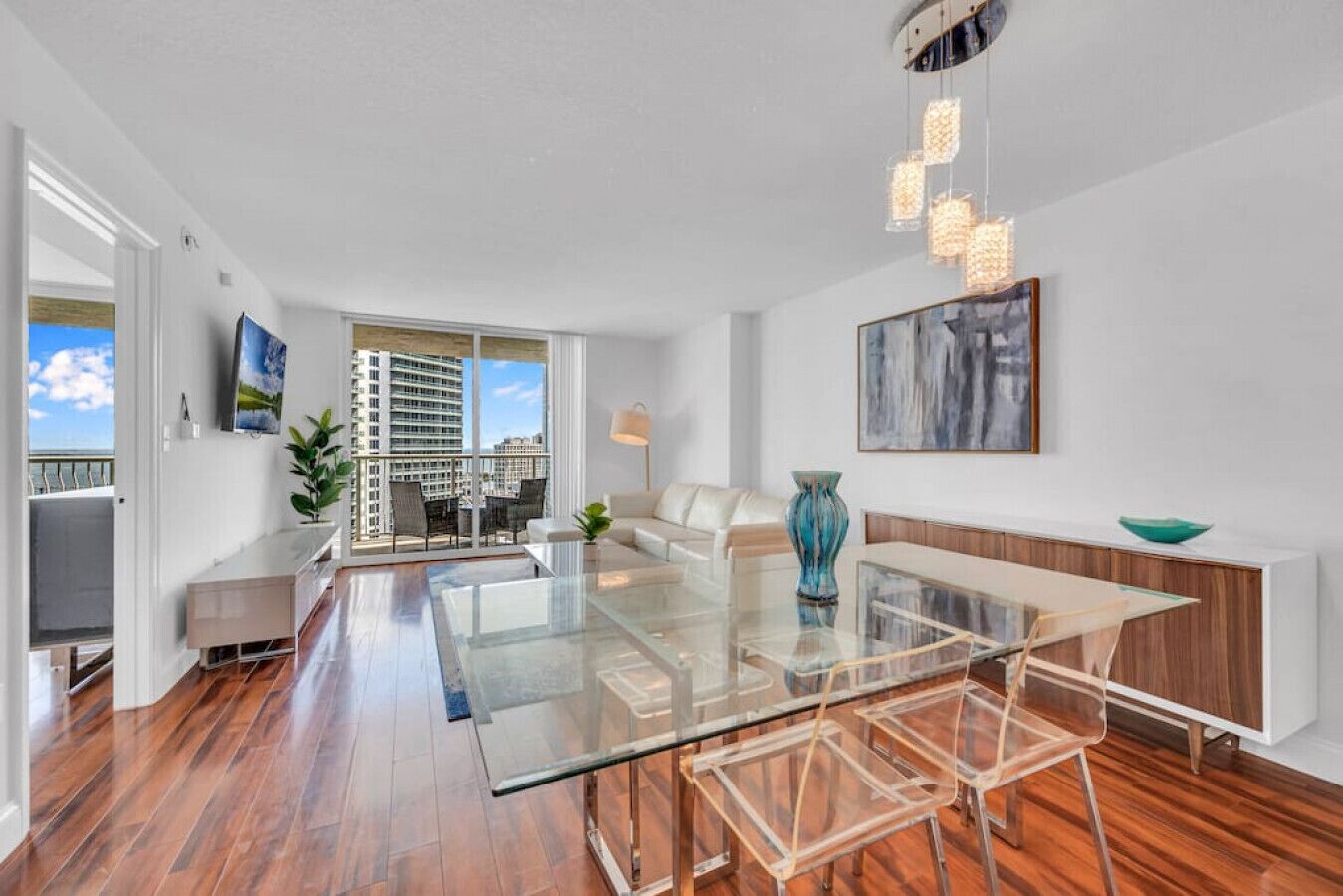 A modern living room with a glass dining table, a sofa, and a view of a cityscape through large windows.
