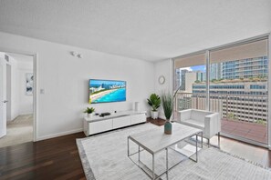 Een moderne woonkamer met een grote televisie, witte meubels, een balkon met uitzicht op de stad en een houten vloer.