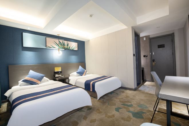 A hotel room with two beds, a desk, a chair, a nightstand, a wall-mounted lamp, and a door.