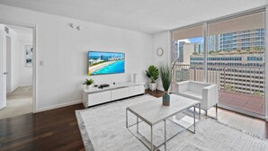 A modern living room with a large TV, white furniture, a balcony with a city view, and a wooden floor.