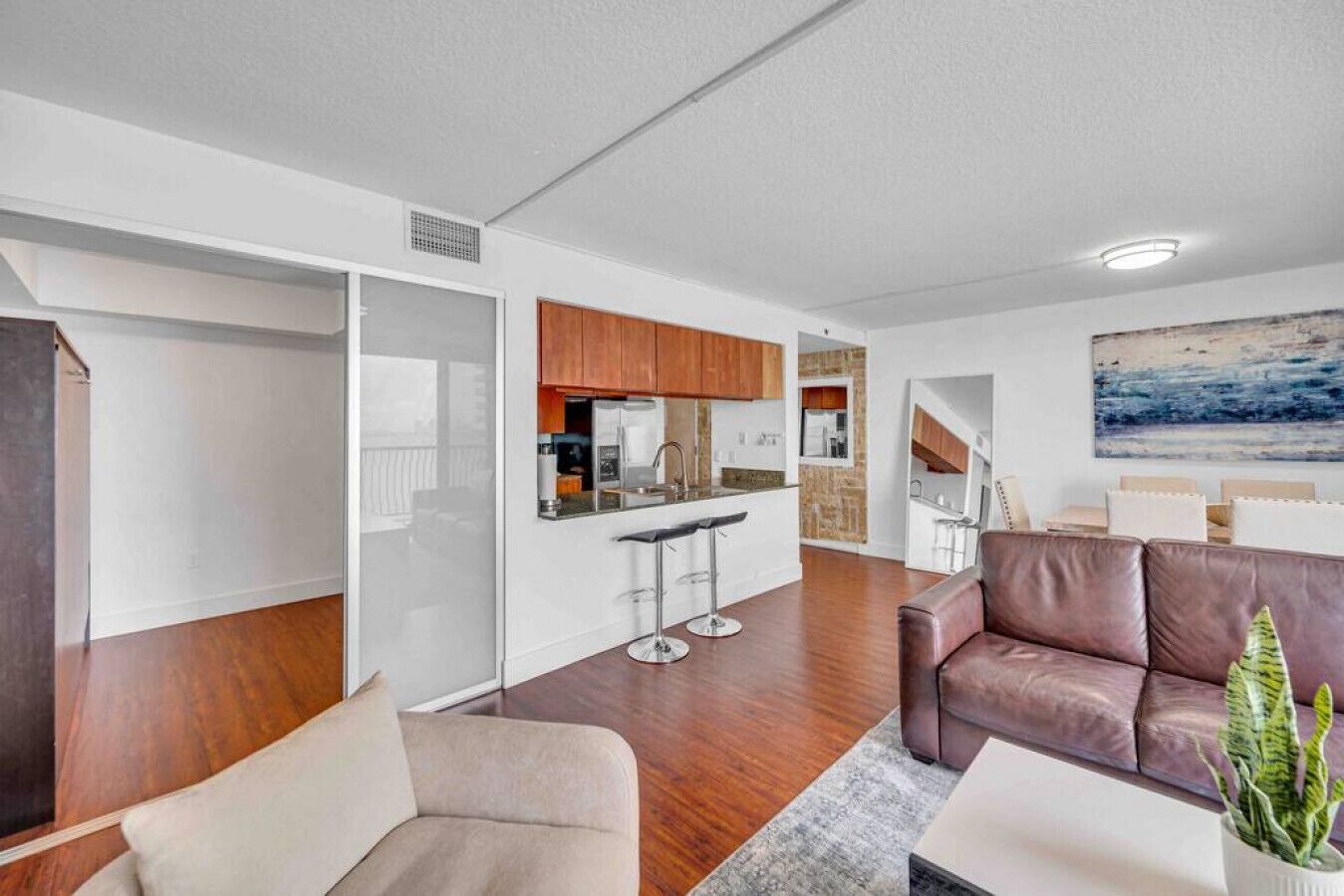 A modern living room with a brown leather sofa, a beige armchair, and a coffee table. A kitchen area with wooden cabinets and a bar stool is visible in the background.