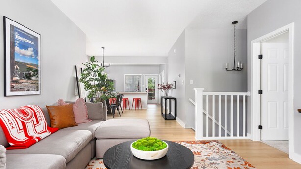 A modern living room with a sectional sofa, a round coffee table with a green centerpiece, and a patterned rug. A framed poster is on the wall, and a staircase is visible in the background. - Little 5 Condo: King Beds | Ping-Pong | Garage (Bloomington)