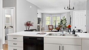 A modern kitchen with a dishwasher, a sink, and a chandelier. There are chairs and a table in the adjacent dining area. - Little 5 Condo: King Beds | Ping-Pong | Garage (Bloomington)