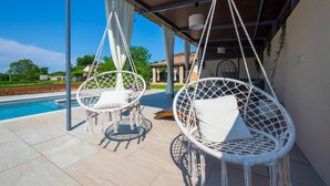 Two white wicker chairs with cushions by a poolside, under a covered area with a view of a house and greenery. - Amazing home in Jadreski with private swimming pool, can be inside or outside (Jadreski)