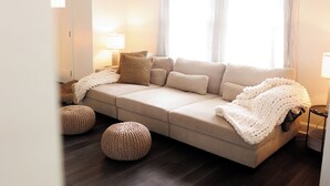 Ein gemütliches Wohnzimmer mit einer Couch, zwei Poufs, einer Lampe und einem Fenster mit Vorhängen.