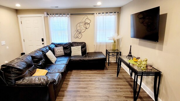 Een woonkamer met een zwarte leren hoekbank, een glazen salontafel en een aan de muur bevestigde televisie.