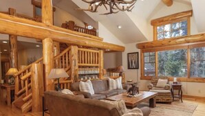 A spacious living room with a wooden staircase, a fireplace, and a large window offering a view of the outdoors. - Amazing Mountain Home in Blue River-7 minutes from Breckenridge! (Blue River)