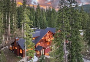 Une grande cabane en rondins comprenant plusieurs pièces, entourée d’une forêt dense et bordée d’un lac.