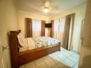 Une chambre avec un lit en bois, des rideaux, un ventilateur de plafond et un sol carrelé.