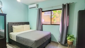 Un dormitorio con cama, aire acondicionado, ventana con cortinas, una planta en maceta y vistas al exterior.