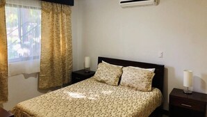 Ein Schlafzimmer mit Bett, Deckenventilator, Klimaanlage, Nachttisch mit Lampe und einem Fenster mit Vorhängen.