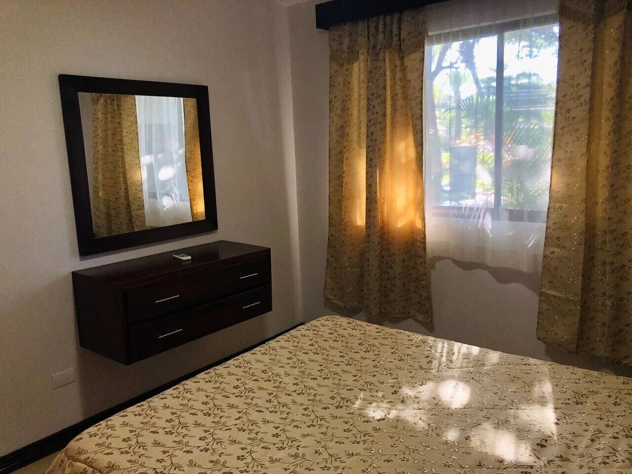 Un dormitorio con cama, cómoda, espejo y ventana con cortinas.