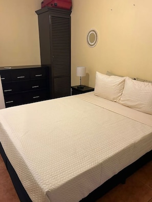 A neatly made bed with a quilted mattress, a bedside table with a lamp, a dark-colored wardrobe, and a wall-mounted round mirror.