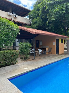 A poolside area with a covered seating area, a pool, and a building with an air conditioning unit.