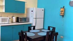 Une cuisine compacte avec des meubles de couleur turquoise, un réfrigérateur blanc, un four à micro-ondes et un petit four. On y trouve une table à manger avec quatre chaises et une horloge murale.