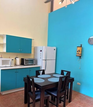 A compact kitchen with teal cabinets, a white refrigerator, a microwave, and a small oven. There is a dining table with four chairs and a wall clock.