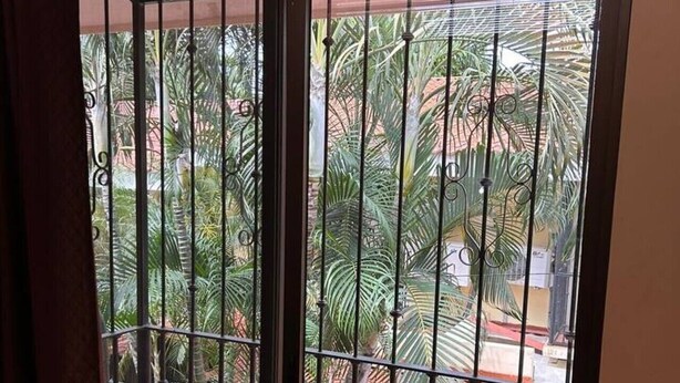 Un balcon avec des balustrades en métal, une vue sur un jardin planté de palmiers et un sol recouvert de carreaux rouges.
