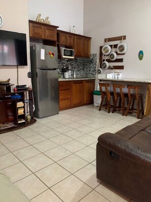 A kitchen with wooden cabinets, a refrigerator, a microwave, a dining table with chairs, and a television.