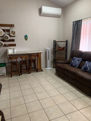 A living room with a brown sofa, a dining area with a table and chairs, and a white air conditioning unit on the wall.