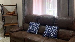 Un canapé en cuir marron agrémenté de deux coussins à motifs, une étagère en bois et une fenêtre avec des rideaux légers.