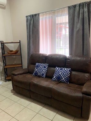 A brown leather sofa with two patterned cushions, a wooden shelf unit, and a window with sheer curtains.