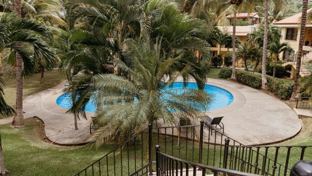 A pool surrounded by palm trees, a concrete deck, and a building with a red roof in the background. - Dolce Vita La Colina in Ocotal (Ocotal)