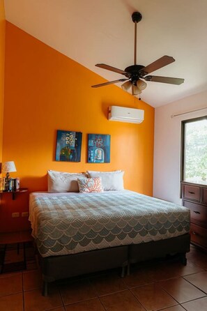 Une chambre à coucher comprenant un lit, un ventilateur de plafond, deux tableaux, un appareil de climatisation, une table de chevet, une lampe et une fenêtre.
