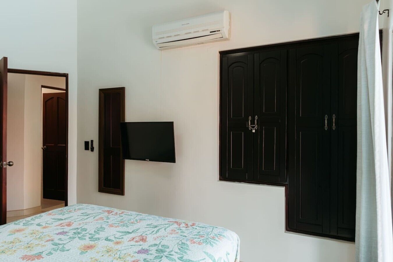 A bedroom with a bed, a TV mounted on the wall, a wardrobe, and an air conditioning unit.