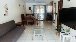A living room with a sofa, a dining table with chairs, a TV, and a kitchen area in the background. - Villa Riviera in Coco Beach (Coco)