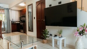 A modern living room with a glass coffee table, a flat-screen TV mounted on the wall, a small white side table, and a potted plant. - Villa Riviera in Coco Beach (Coco)