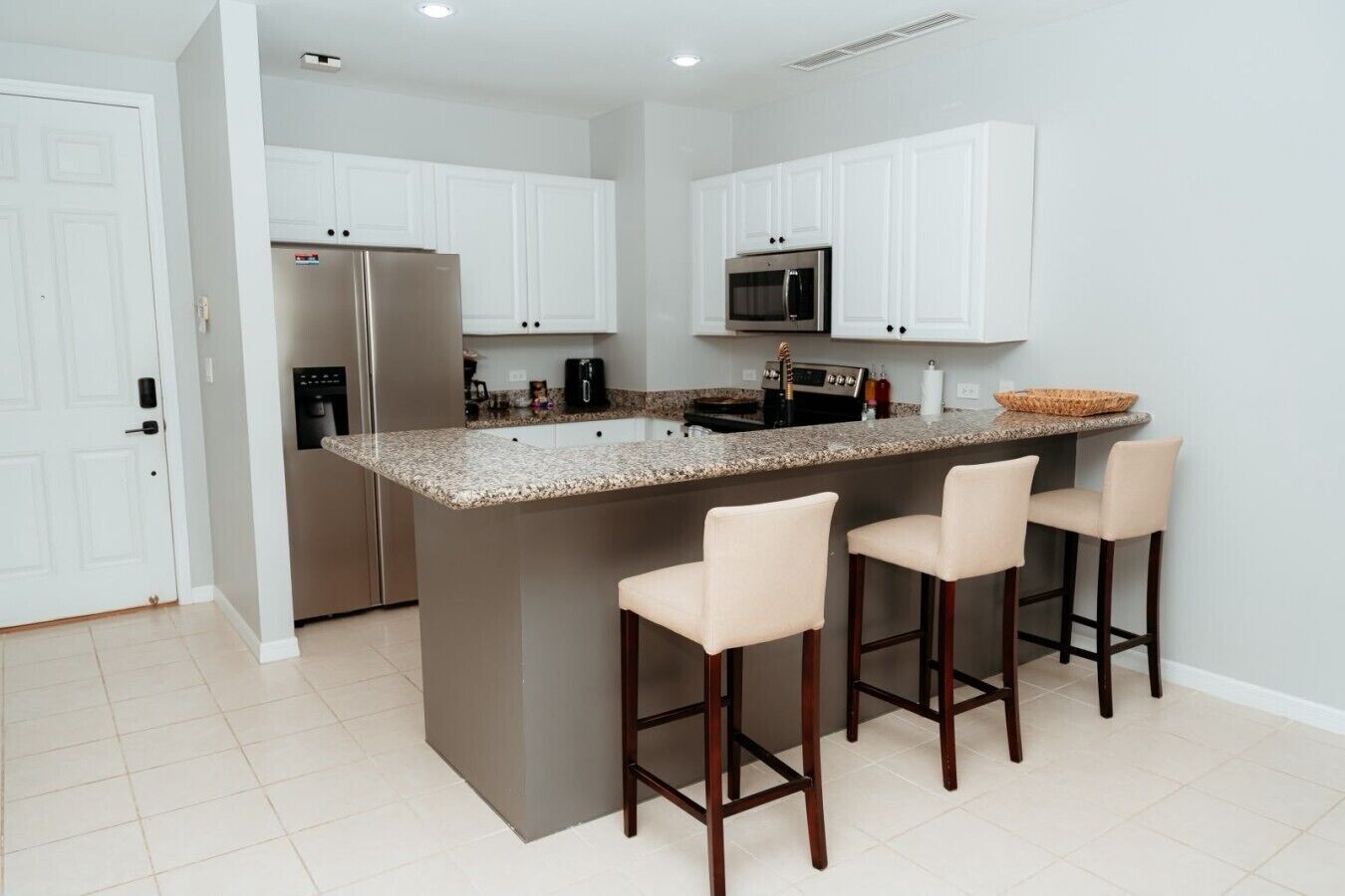 Cocina con gabinetes blancos, refrigerador de acero inoxidable, microondas y una isla de encimera de granito con taburetes de bar.