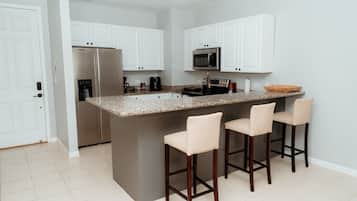 Cocina con gabinetes blancos, refrigerador de acero inoxidable, microondas y una isla de encimera de granito con taburetes de bar.