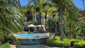 A swimming pool with a stone edge, surrounded by lush greenery and palm trees. - Lovely 2 bdrm Condo in Pacifico (Coco)